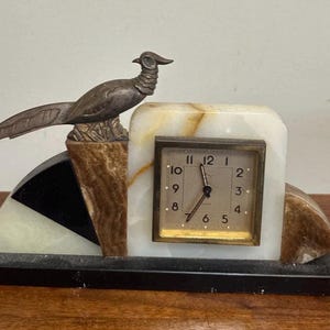 May include: A mantel clock featuring a metallic pheasant figurine perched on a multi-colored stone base. The clock has a square face with black numerals and hands, set into a white, brown, and black stone structure with a rectangular black base.
