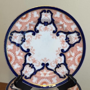 May include: A stack of decorative plates with a star-shaped design. Each plate features a white center, a blue border with gold trim, and a pattern of orange floral motifs. The plates are displayed on black stands.