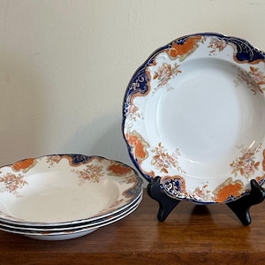 Puede incluir: Un juego de cuatro cuencos de sopa de cerámica blanca con borde festoneado y una cenefa decorativa en azul marino intenso y naranja quemado, complementado con intrincados motivos florales en tonos rosa y verde en el borde.