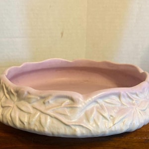 Pode incluir: Uma tigela de cerâmica rosa claro com uma borda ondulada e um design em relevo semelhante a folhas ao redor do exterior. A tigela está sobre uma superfície de madeira.