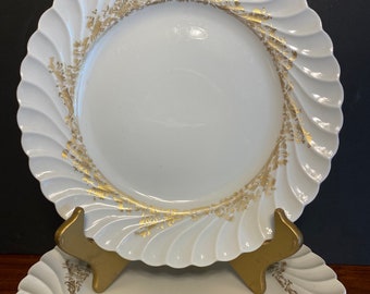 May include: A set of white ceramic plates featuring a scalloped, fluted edge design. Each plate is decorated with a delicate, intricate gold filigree pattern that encircles the inner rim, providing a metallic contrast to the smooth white surface.