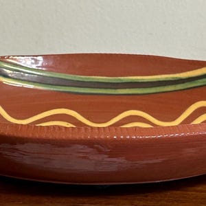 May include: A shallow, oval-shaped ceramic bowl with a glossy terracotta-colored glaze. The interior features decorative hand-painted wavy lines in cream and dark green, with a textured, ribbed pattern along the outer rim of the bowl.