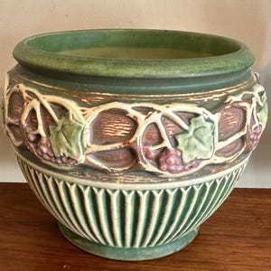 May include: A green and brown ceramic planter with a decorative grape vine design. The planter has a wide, rounded shape with a ribbed base and a textured rim. The grape vine design features clusters of purple grapes and green leaves.