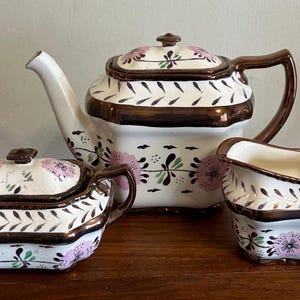 May include: A ceramic tea set consisting of a teapot with a lid, a lidded sugar bowl, and a creamer. Each piece is white with a metallic brown trim, featuring a repeating pattern of pink flowers and dark green leaf motifs on the exterior.