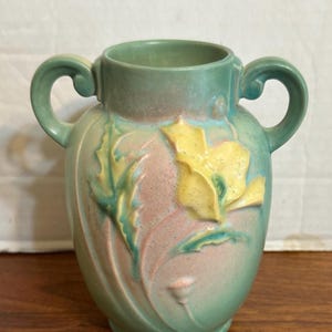 Puede incluir: Un jarrón de cerámica vintage en tonos verdes, rosas y amarillos. El jarrón presenta dos asas curvas y un diseño floral con una flor amarilla y hojas verdes. El jarrón mide aproximadamente 18 cm de alto.