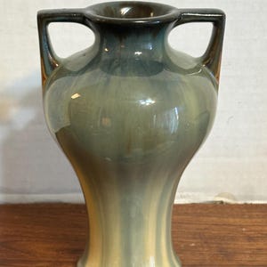 May include: A ceramic vase with two handles, featuring a gradient of colors from dark brown to light green and cream. The vase has a classic shape with a wide base and a narrow neck.