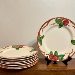 May include: A set of cream-colored ceramic plates with a brown rim and a decorative apple and leaf design. The plates are stacked and displayed on a wooden surface. The plates are a classic style.