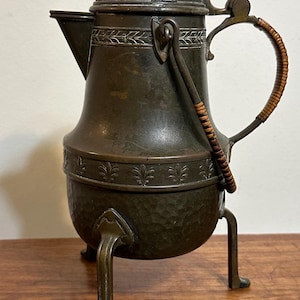 Könnte beinhalten: Ein dunkel bronzefarbener Metallkrug mit Klappdeckel, Ausguss und drei Beinen. Der Körper weist im unteren Bereich eine gehämmerte Textur auf, während das obere Band mit geprägten Blattmustern verziert ist. Der Metallgriff ist mit Rohr umwickelt.