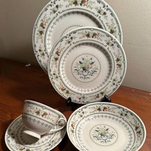 Op de afbeelding: Een verzameling wit keramisch servies met een bloemenontwerp. De set bevat borden, een kom en een theekopje met schotel. De randen en centra hebben een groen en bloemenpatroon.