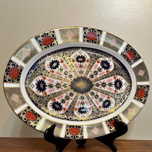 May include: An oval porcelain serving platter featuring a central medallion with radiating floral patterns in navy blue, white, and gold. The wide rim is decorated with alternating panels of red flowers, gold lattice, and navy blue geometric motifs.