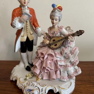 May include: A porcelain figurine depicting a man and woman in 18th-century attire. The man wears an orange coat and holds a paper, while the woman plays a mandolin. The base is white with gold accents and floral details. The woman's dress is pink and white.