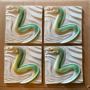 Puede incluir: Cuatro baldosas de cerámica cuadradas, cada una con una escultura en relieve de una anguila estilizada. Las anguilas son de color verde y amarillo, sobre un fondo texturizado con forma de olas. Disposición en cuadrícula 2x2.
