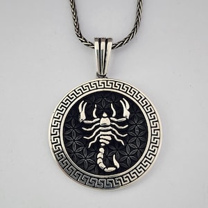 May include: A silver pendant necklace with a circular pendant featuring a detailed scorpion design. The pendant has a textured background and a Greek key border. The chain is a woven style.