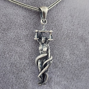 May include: A silver pendant necklace featuring a detailed depiction of Lady Justice, blindfolded and holding scales and a sword, with a snake wrapped around her body.