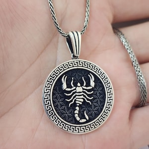 May include: A silver pendant necklace with a circular pendant depicting a scorpion. The scorpion is white against a black, floral-patterned background. The pendant has a Greek key border and is suspended from a braided chain.