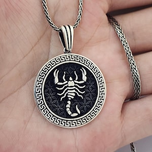 May include: A silver pendant necklace with a detailed scorpion design within a circular frame. The pendant has a black background with a floral pattern and a Greek key border. The chain is a woven style.