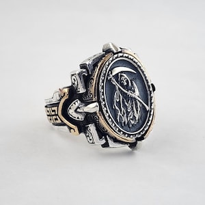 May include: A silver and gold ring with a detailed design featuring a grim reaper holding a scythe. The ring has a black enamel inlay with a detailed image of the grim reaper.