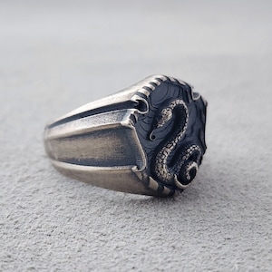May include: A silver ring featuring a detailed snake design. The ring has a textured, ridged band and a raised snake design on the top.