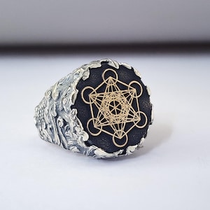 May include: A silver ring with a black and gold geometric design. The ring has a textured band and a round top.