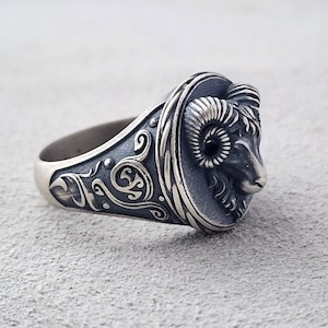 Może przedstawiać: Srebrny pierścionek z detalicznym wzorem głowy barana. Pasek pierścionka jest ozdobiony misternymi wzorami kwiatowymi.