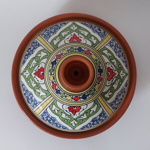 May include: A terracotta tagine with a conical lid featuring a central handle. The lid is decorated with a symmetrical pattern in blue, green, yellow, and red, arranged in four distinct quadrants around a circular center with geometric shapes.