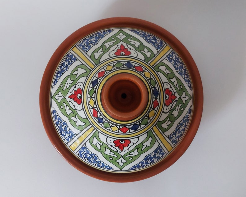 May include: A terracotta tagine with a conical lid featuring a central handle. The lid is decorated with a symmetrical pattern in blue, green, yellow, and red, arranged in four distinct quadrants around a circular center with geometric shapes.