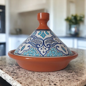 May include: A terracotta tagine featuring a conical lid with a hand-painted blue, turquoise, and white floral pattern. The base is a wide, shallow, unglazed reddish-brown clay bowl with a smooth rim, topped by a matching pointed lid handle.