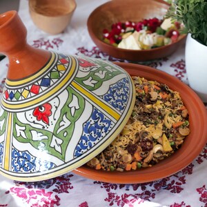May include: A terracotta tagine with a conical lid featuring hand-painted geometric and floral patterns in blue, green, red, and yellow. The base contains a cooked dish of noodles, mushrooms, carrots, and peas, partially covered by the lid.