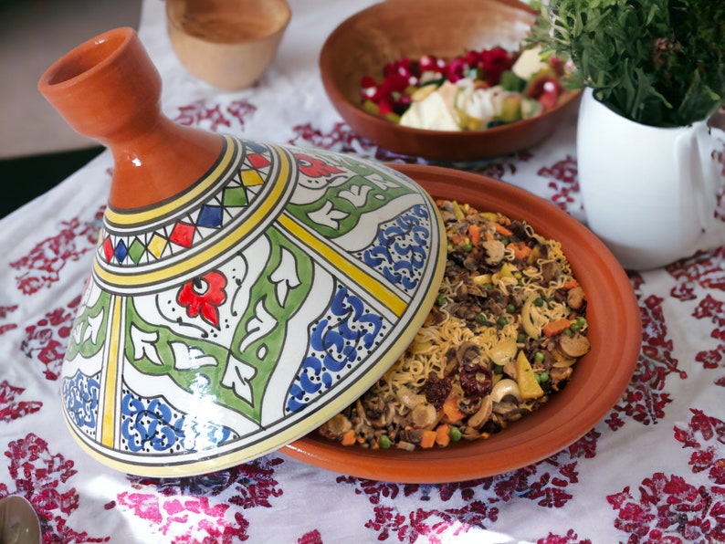 May include: A terracotta tagine with a conical lid featuring hand-painted geometric and floral patterns in blue, green, red, and yellow. The base contains a cooked dish of noodles, mushrooms, carrots, and peas, partially covered by the lid.