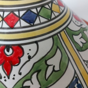 May include: A ceramic tajine lid featuring a conical shape with a white base and hand-painted details. The surface displays vibrant red, green, blue, and yellow floral motifs, geometric diamond patterns, and black outlines on a glazed finish.