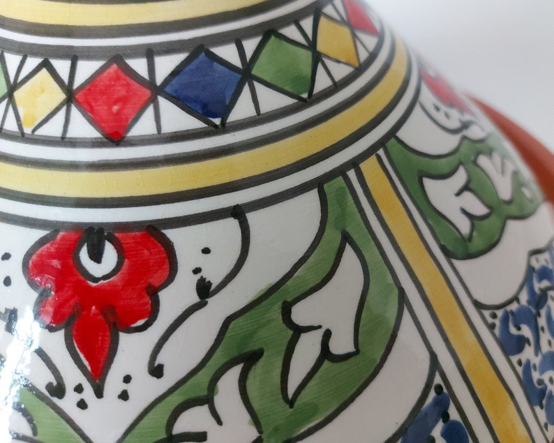 May include: A ceramic tajine lid featuring a conical shape with a white base and hand-painted details. The surface displays vibrant red, green, blue, and yellow floral motifs, geometric diamond patterns, and black outlines on a glazed finish.