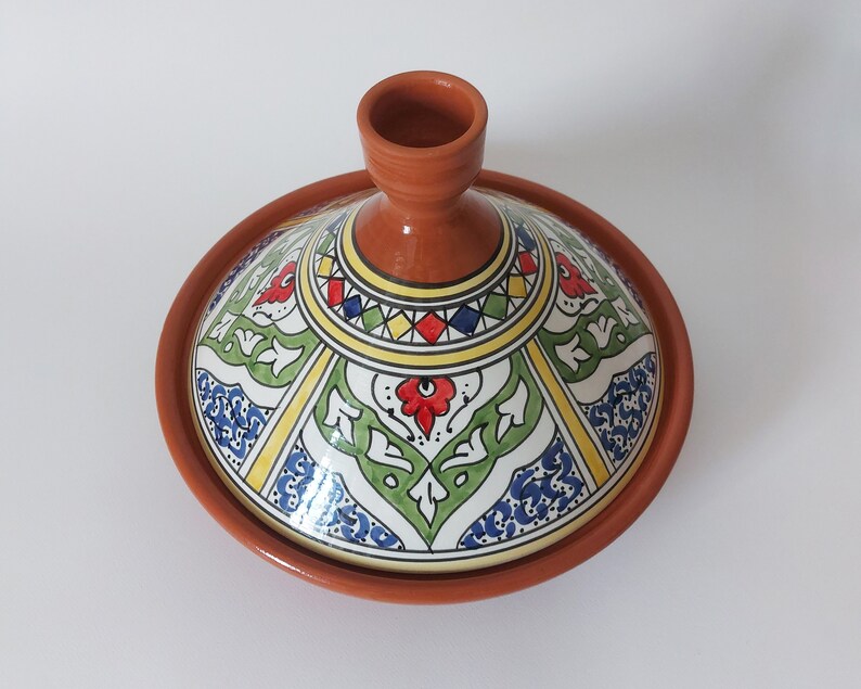 May include: A terracotta tagine with a conical lid featuring hand-painted geometric and floral patterns in red, green, blue, and yellow. The unglazed clay base contrasts with the glossy, multi-colored glazed lid, which has a central knob handle.