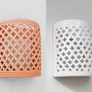 Puede incluir: Dos apliques de pared semicilíndricos, uno en color terracota naranja mate y el otro con acabado blanco brillante. Ambos accesorios presentan un patrón de celosía repetitivo de formas de corazón recortadas en la parte frontal, permitiendo el paso de luz.