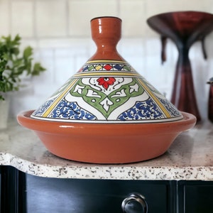 May include: A terracotta tagine featuring a conical lid with a hand-painted design in green, blue, red, and yellow. The base is a wide, shallow bowl with a smooth, unglazed finish. The lid has a central knob handle and intricate floral patterns.