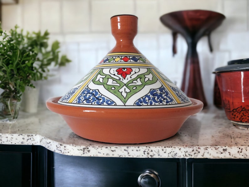 May include: A terracotta tagine featuring a conical lid with a hand-painted design in green, blue, red, and yellow. The base is a wide, shallow bowl with a smooth, unglazed finish. The lid has a central knob handle and intricate floral patterns.
