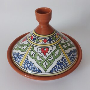 May include: A terracotta tagine with a conical lid featuring a hand-painted design. The lid displays a geometric pattern of red, blue, green, and yellow shapes, along with floral motifs, set against a white background with a smooth finish.