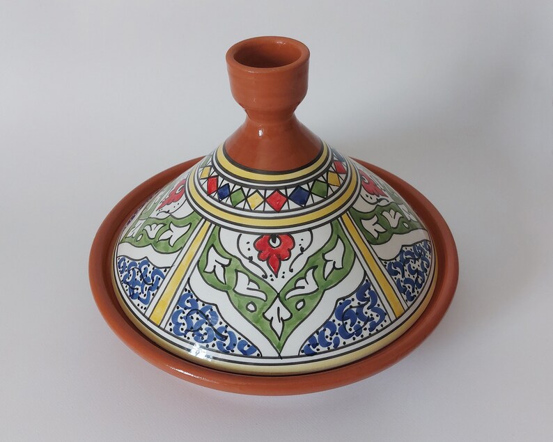 May include: A terracotta tagine with a conical lid featuring a hand-painted design. The lid displays a geometric pattern of red, blue, green, and yellow shapes, along with floral motifs, set against a white background with a smooth finish.