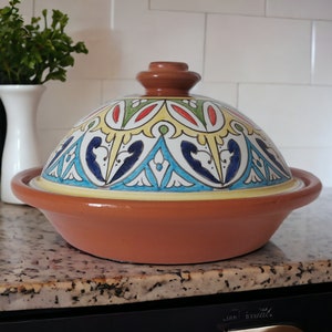 May include: A terracotta tagine with a conical lid featuring hand-painted geometric and floral patterns in blue, yellow, green, and red. The base is a wide, shallow, unglazed brown clay bowl with a matching brown knob handle on the lid top.