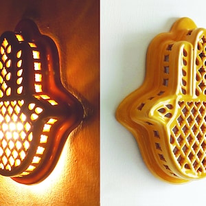 May include: A yellow ceramic wall lamp shaped like a hamsa hand with intricate cut-out lattice patterns. The lamp is shown in two states: illuminated with a warm glow emitting through the openings and unlit against a plain white background.