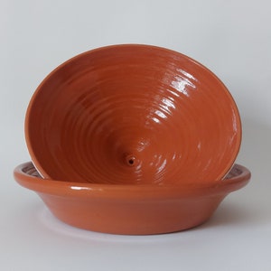 May include: A terracotta-colored ceramic bowl with a wide, flat base and a deep, conical interior featuring a small central hole. The surface has a smooth, glossy finish with visible concentric circular ridges throughout the interior basin.