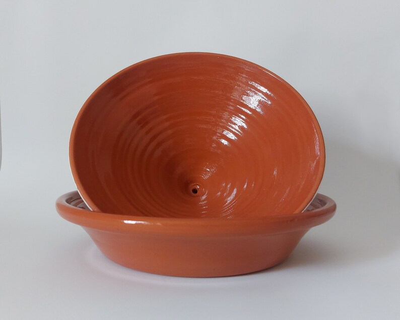 May include: A terracotta-colored ceramic bowl with a wide, flat base and a deep, conical interior featuring a small central hole. The surface has a smooth, glossy finish with visible concentric circular ridges throughout the interior basin.