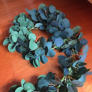 Puede incluir: Una larga guirlanda de hojas de eucalipto artificiales en tonos verdes. Las hojas están dispuestas en cascada, creando un aspecto exuberante y natural.