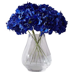 Puede incluir: Un ramo de hortensias azules en un jarrón de vidrio transparente. El jarrón tiene una base redonda y una abertura ancha.