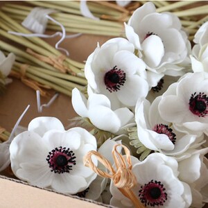 May include: A bunch of white artificial anemones with dark centers in a cardboard box. The flowers have green stems and are tied together with twine.