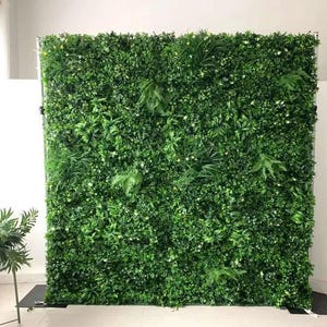 May include: A large artificial green wall, featuring a variety of faux plants and foliage. The wall is rectangular and stands upright, creating a backdrop. The plants are a mix of textures and shades of green, with some small white flowers.