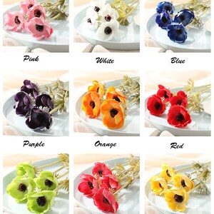 May include: A set of 10 artificial anemone flower stems in various colors, including pink, white, blue, purple, orange, red, green, fuchsia, yellow. The flowers are arranged in a bouquet on a white plate.