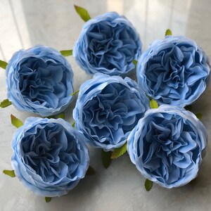 Może przedstawiać: Sześć jasnoniebieskich sztucznych głów róż z zielonymi łodygami. Kwiaty są ułożone w kółko na białym tle.