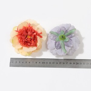 Puede incluir: Dos cabezas de flores artificiales, una es una peon&iacute;a de color melocot&oacute;n y crema con un centro rojo, la otra es una flor de color p&uacute;rpura claro con un centro verde.