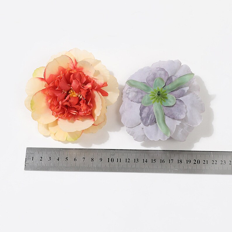 Puede incluir: Dos cabezas de flores artificiales, una es una peon&iacute;a de color melocot&oacute;n y crema con un centro rojo, la otra es una flor de color p&uacute;rpura claro con un centro verde.