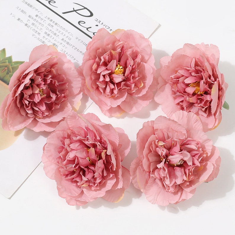Puede incluir: Cinco flores de peon&iacute;a artificiales de color rosa con un aspecto en capas y plisado. Las flores est&aacute;n dispuestas sobre una superficie blanca.